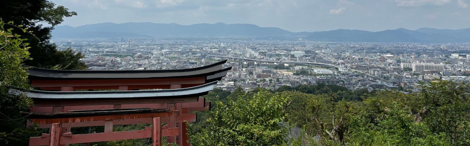 A red torii gate stands in the foreground overlooking a city surrounded by mountains. 